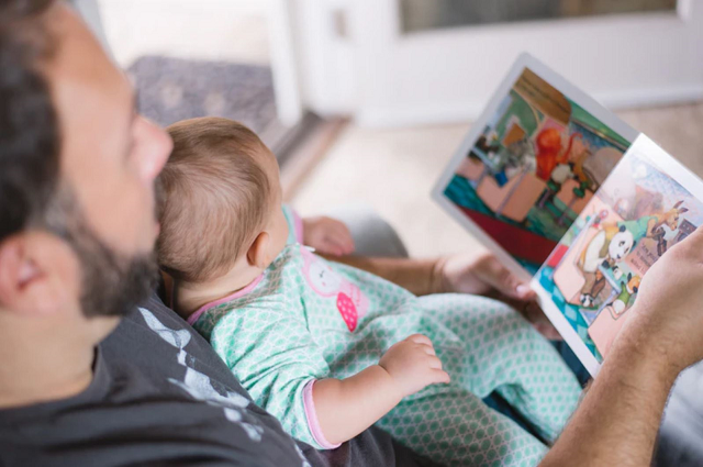 30 libros para bebé que deberás tener en tu lista de regalos, a father sitting on the couch reading a book to his baby, who is leaning against him.